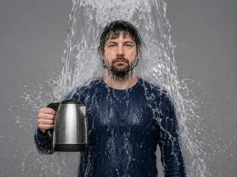 Man in a blue long-sleeved shirt holding a kettle while drenched with water against a grey background, illustrating the need to rinse thoroughly after descaling.